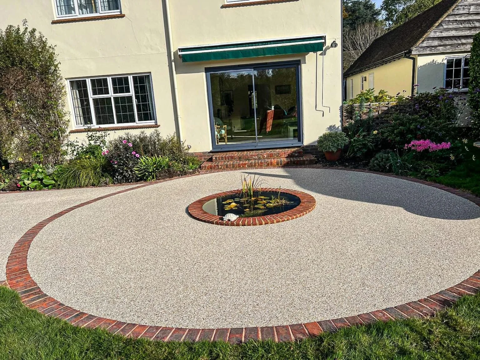 Backyard with a small pond in the center surrounded by a gravel patio area edged with bricks, mother separates a flower bed with colorful flowers and bushes, with a house and windows in the background.