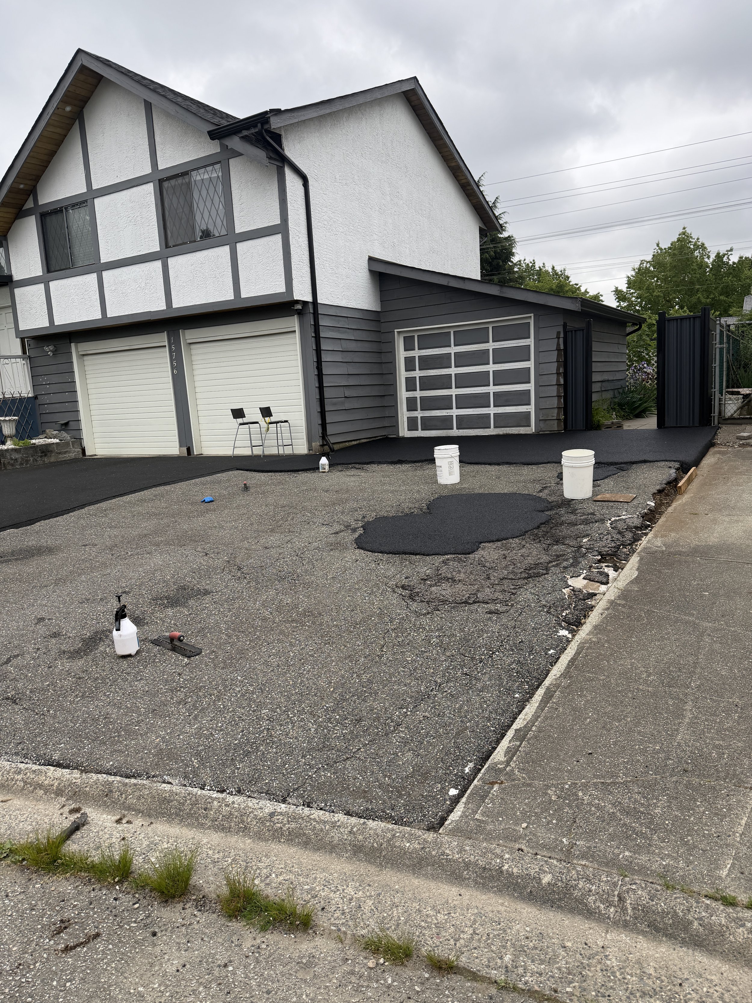 A house with a black and white exterior, garage doors, and a driveway that is being resurfaced with fresh asphalt. Construction supplies and tools are visible, including a spray bottle, buckets, and roller.
