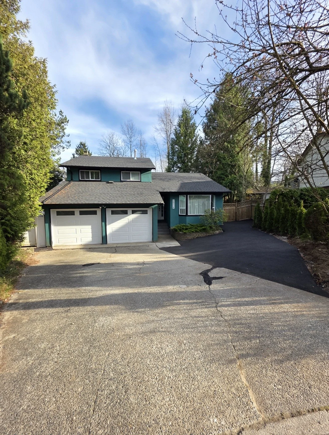 A two-story house with teal exterior walls, black roof, white garage doors, a large front window with white curtains, and a small garden bed in front. The driveway is paved in black asphalt, leading to the garage, with an expanding parking area in front. Trees with no leaves and partly cloudy sky are in the background.