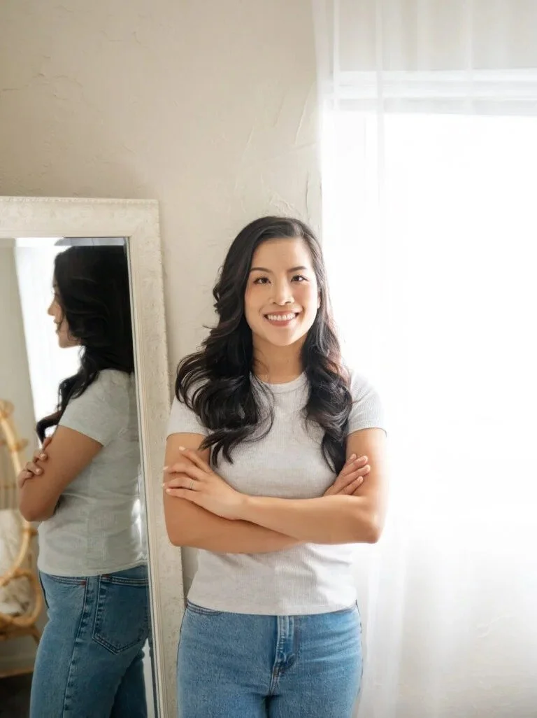 A young woman with long dark hair smiling and crossing her arms, standing near a mirror in a well-lit room.
