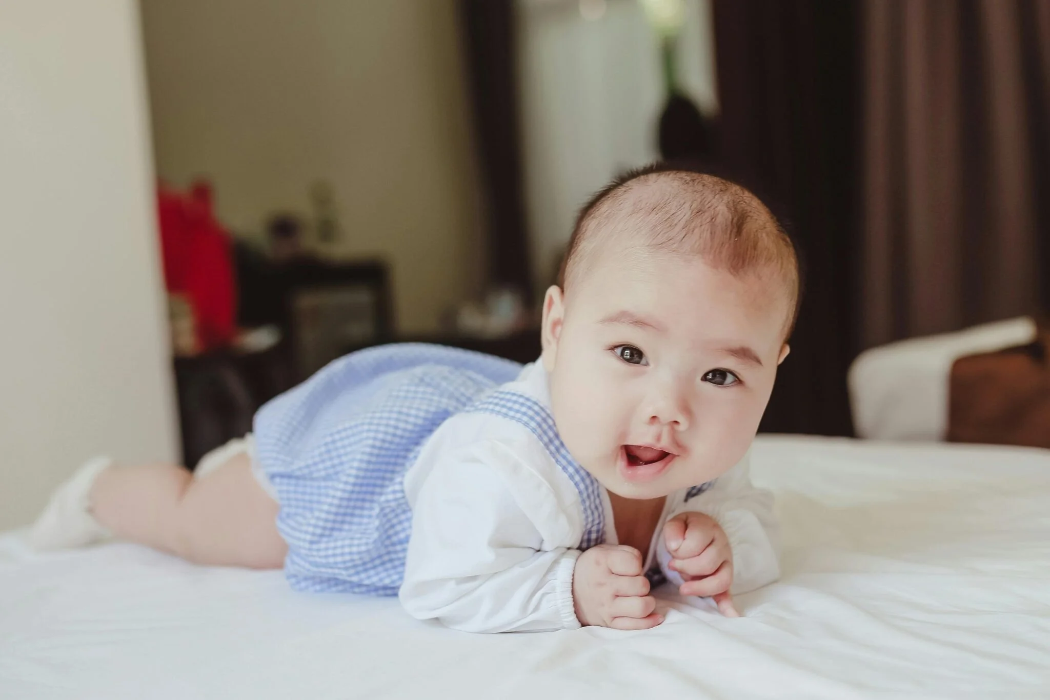 A baby lying on their stomach on a bed, looking at the camera with their hands clasped. The baby has a bald head and is wearing a white long-sleeve shirt with a blue checkered vest. The background is slightly blurred with dark curtains and some furniture.