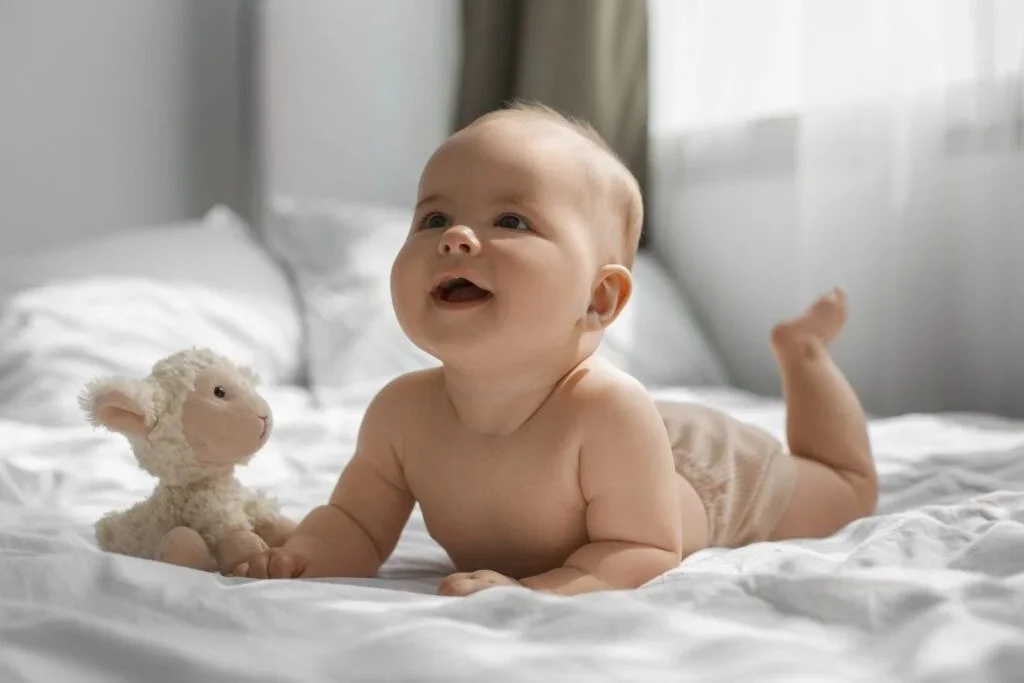 A smiling baby lying on a bed with white sheets, looking up, with a small plush lamb toy beside them.