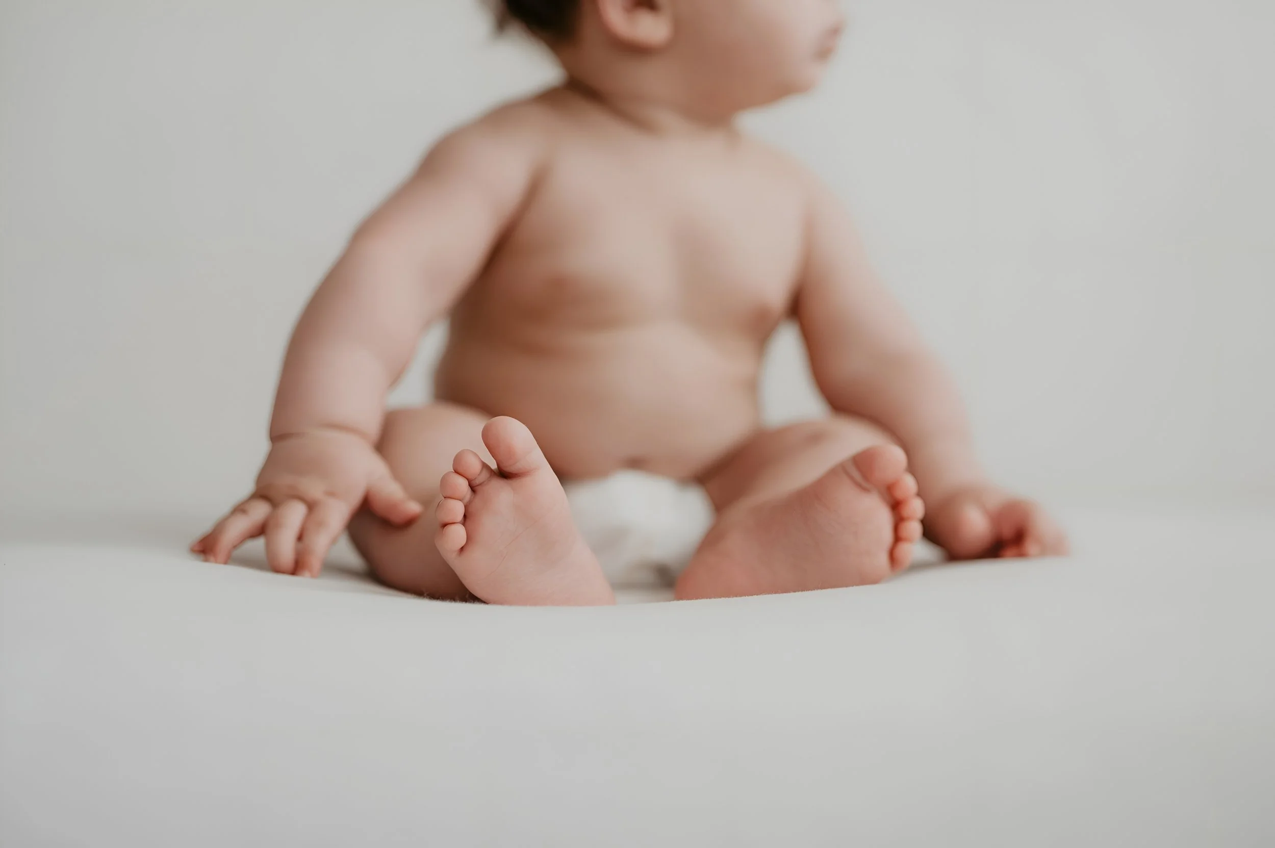 A close-up of a baby sitting on a white surface, with visible chubby arms, legs, and feet, and wearing only a diaper.