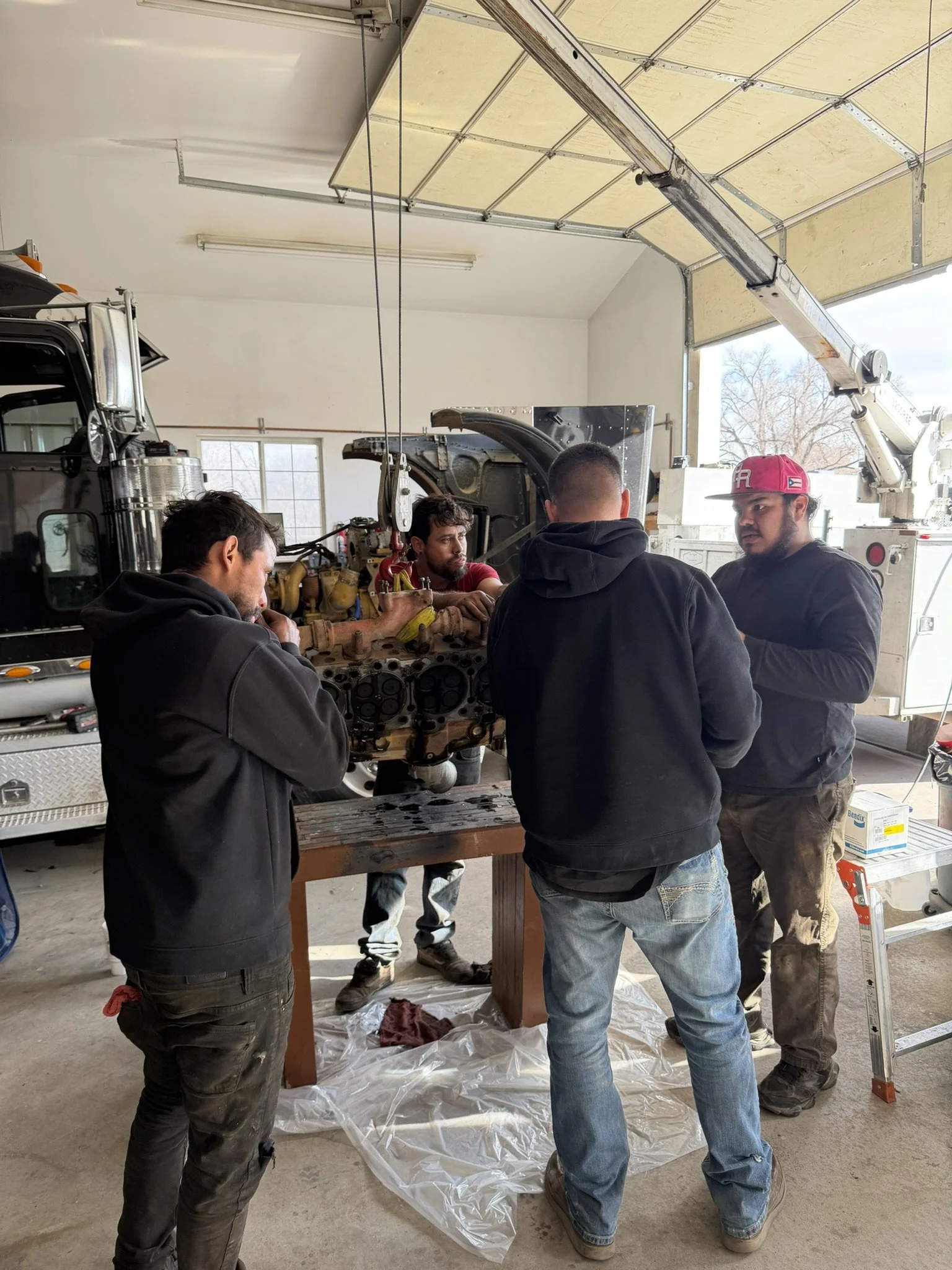 Four men are working on an engine in a garage or workshop, with a large vehicle or truck nearby and a crane or boom in the background.