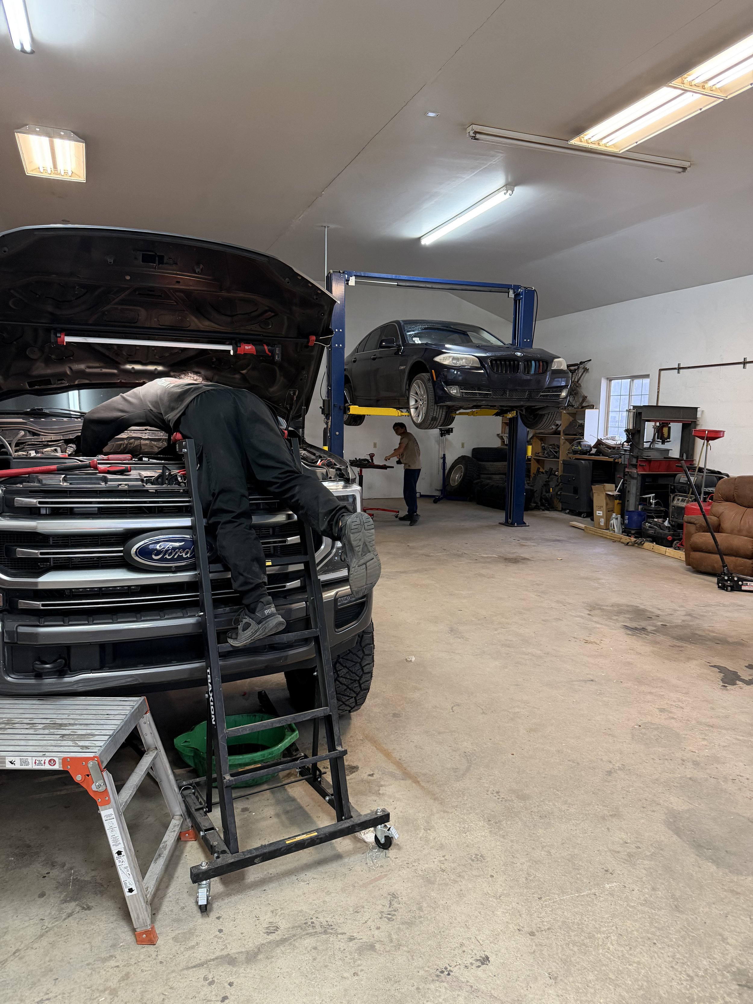 A mechanic working under the hood of a black Ford truck in a garage, with another car elevated on a lift and a person working in the background.