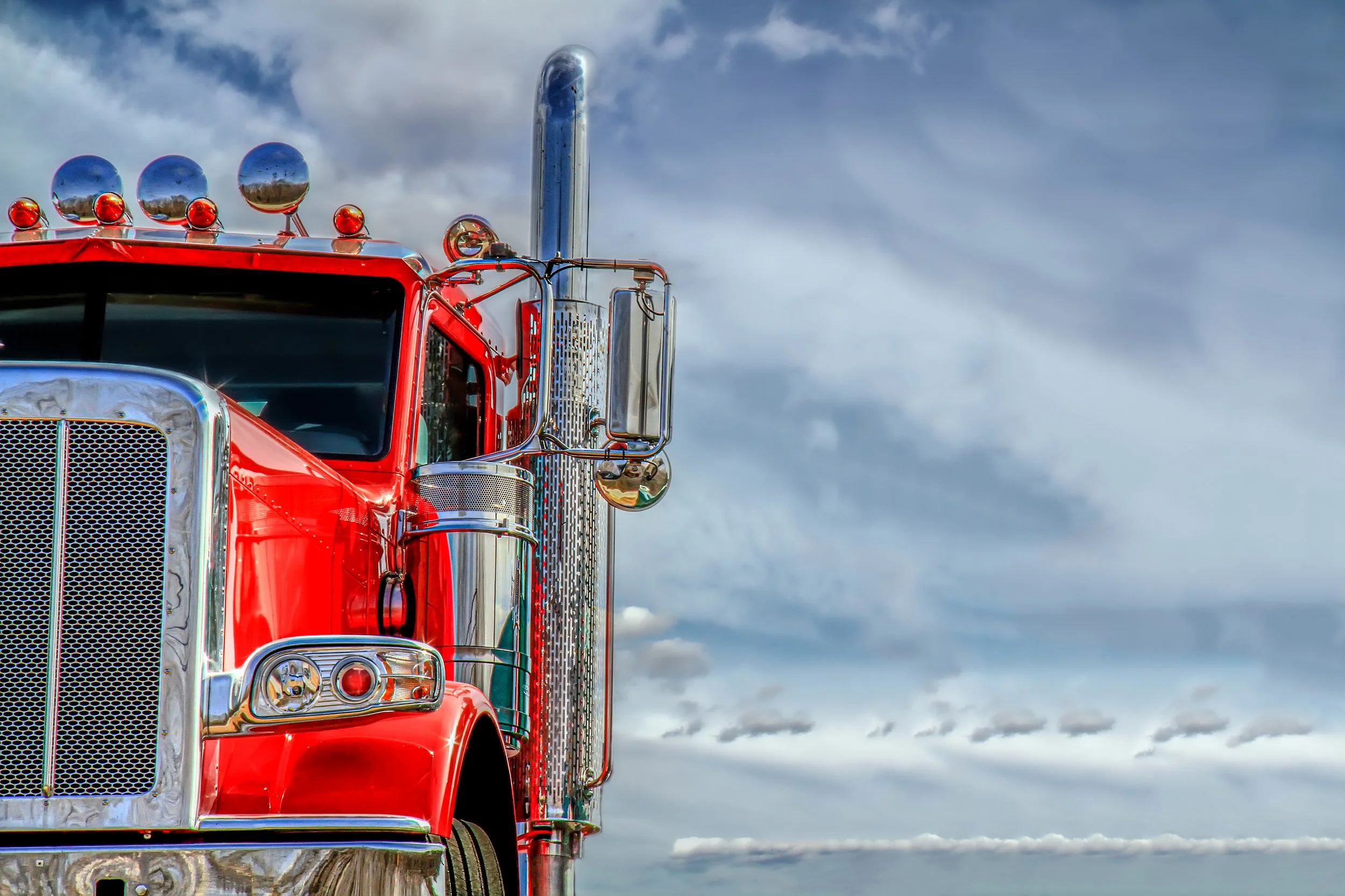 Front half of a red semi-truck with chrome details and a high exhaust pipe, set against a background of cloudy sky.