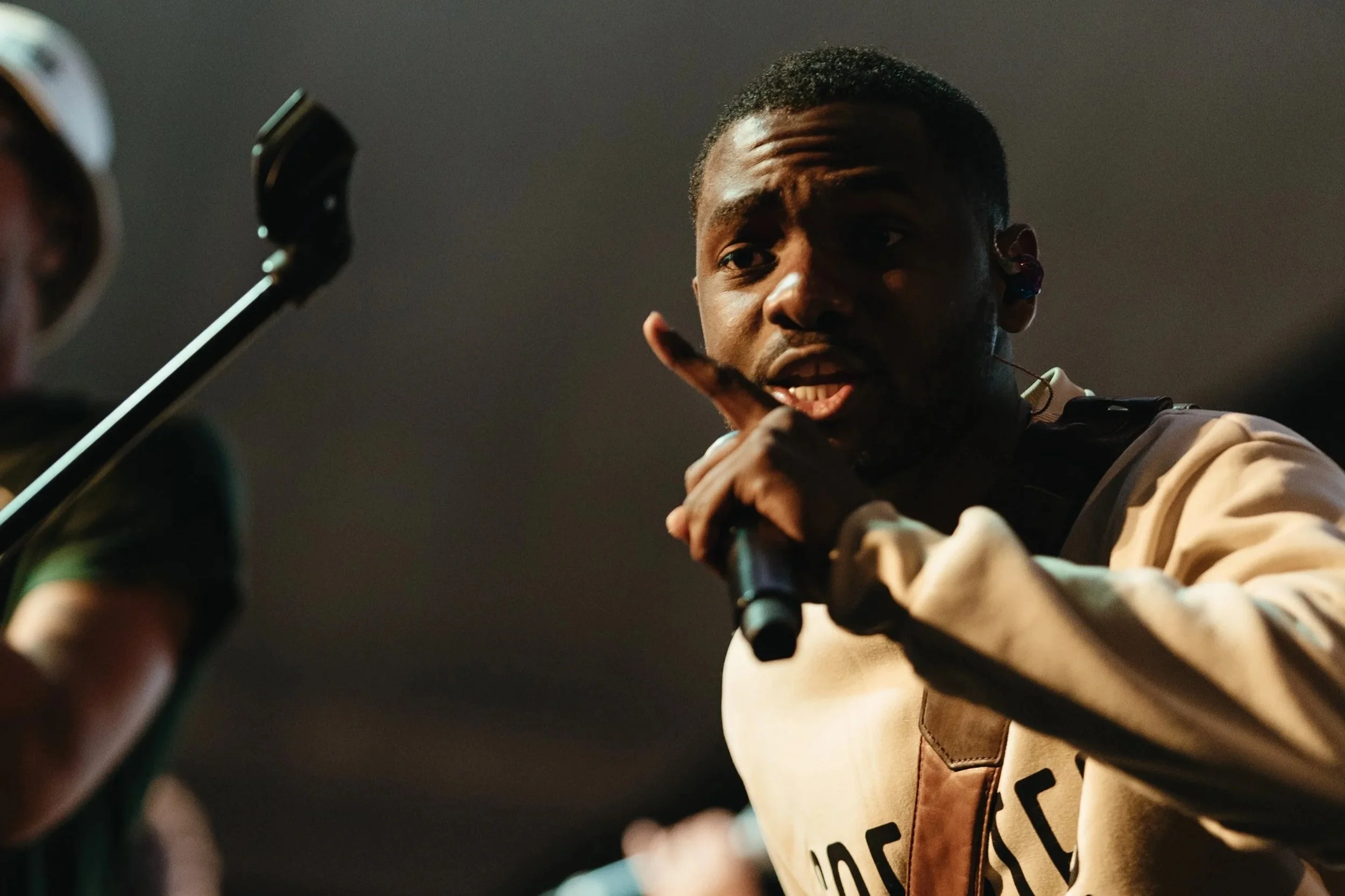 A man in a beige and black jacket holding a microphone, speaking or singing with a serious expression, while another person with a microphone stands nearby.