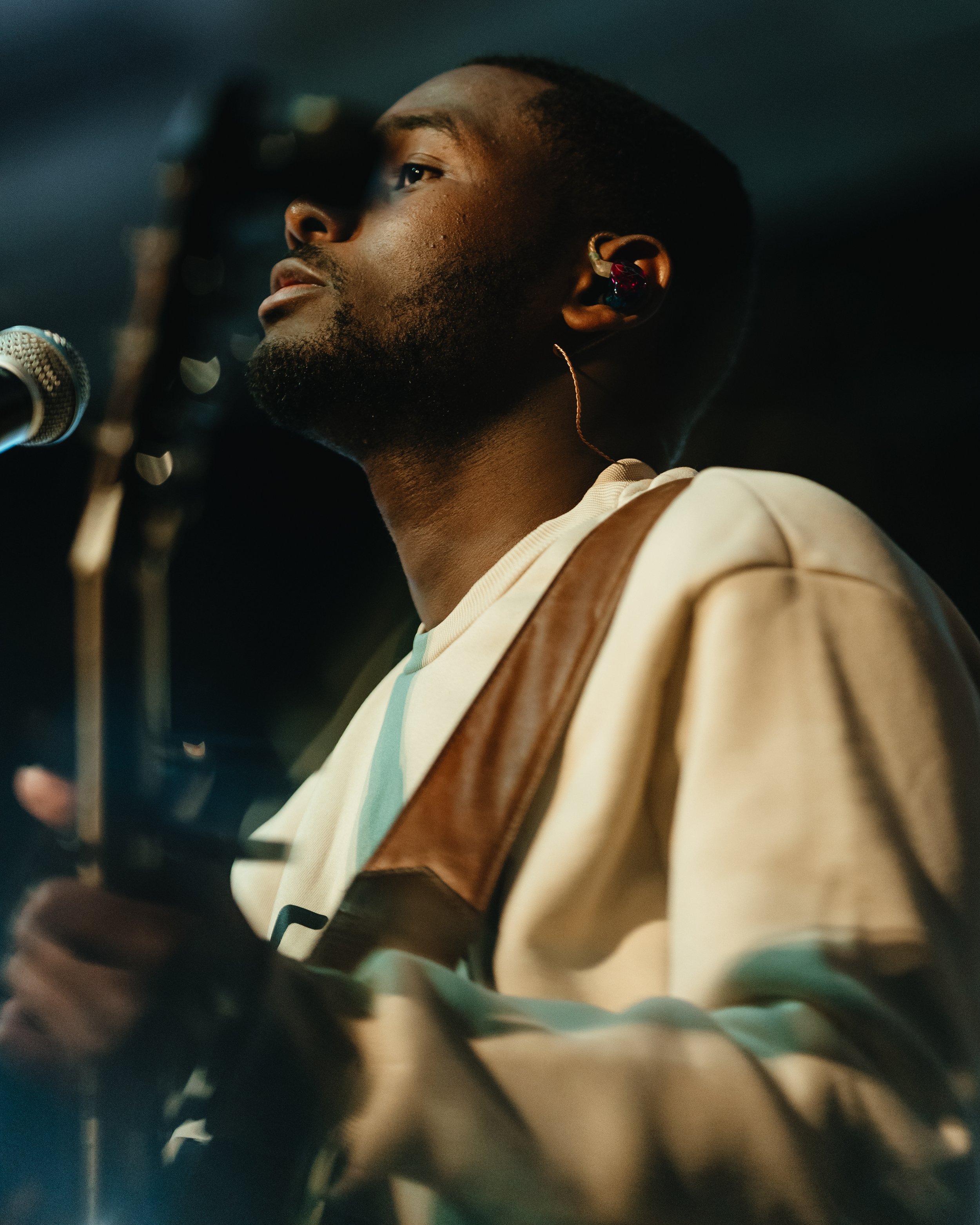 A young man with short hair and a beard, wearing a beige sweatshirt, playing a guitar and singing into a microphone. He has in-ear monitors and is focused on his performance.