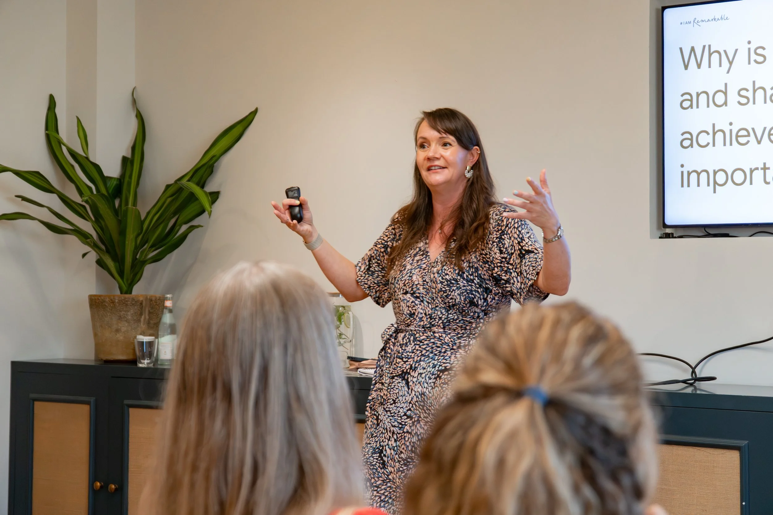 A woman giving a presentation in a room with an audience. She is holding a remote and gesturing with her other hand. A plant is in the corner and a screen displays text about achievement and importance.