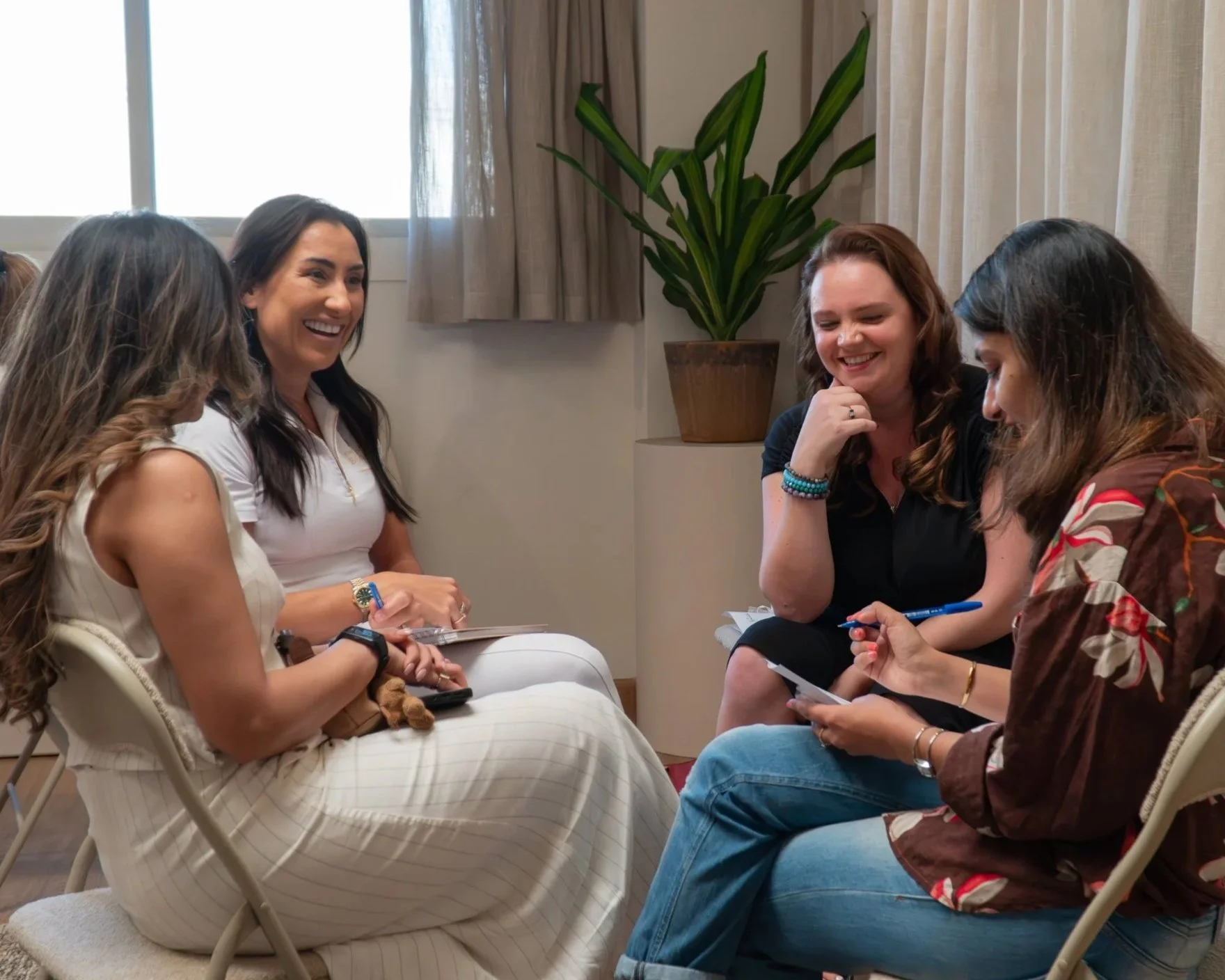 Four women sitting in a circle, laughing and chatting during a casual meeting in a bright room with a plant and window in the background.