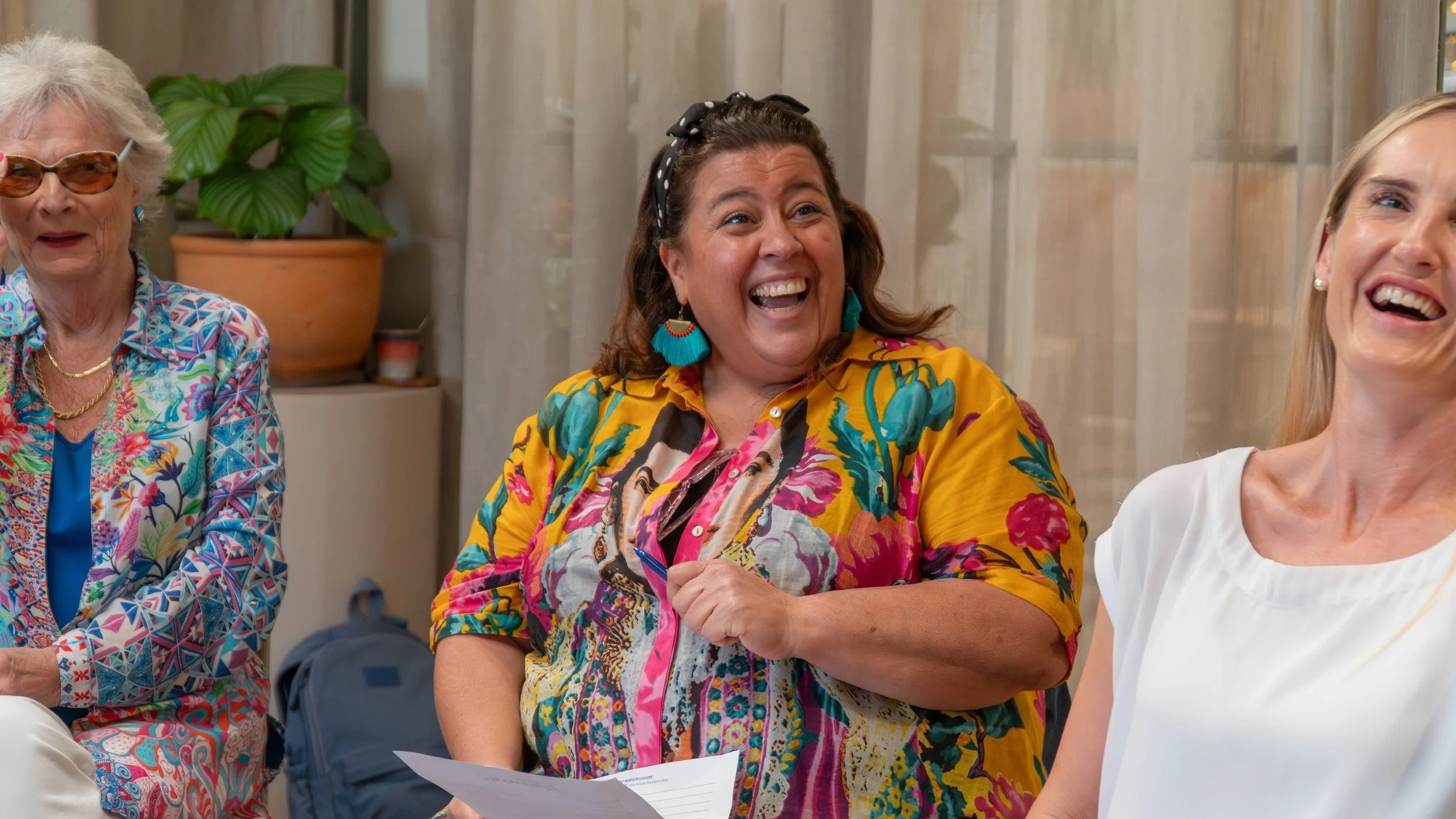 Three women laughing indoors, with a potted plant and light-colored curtains in the background.