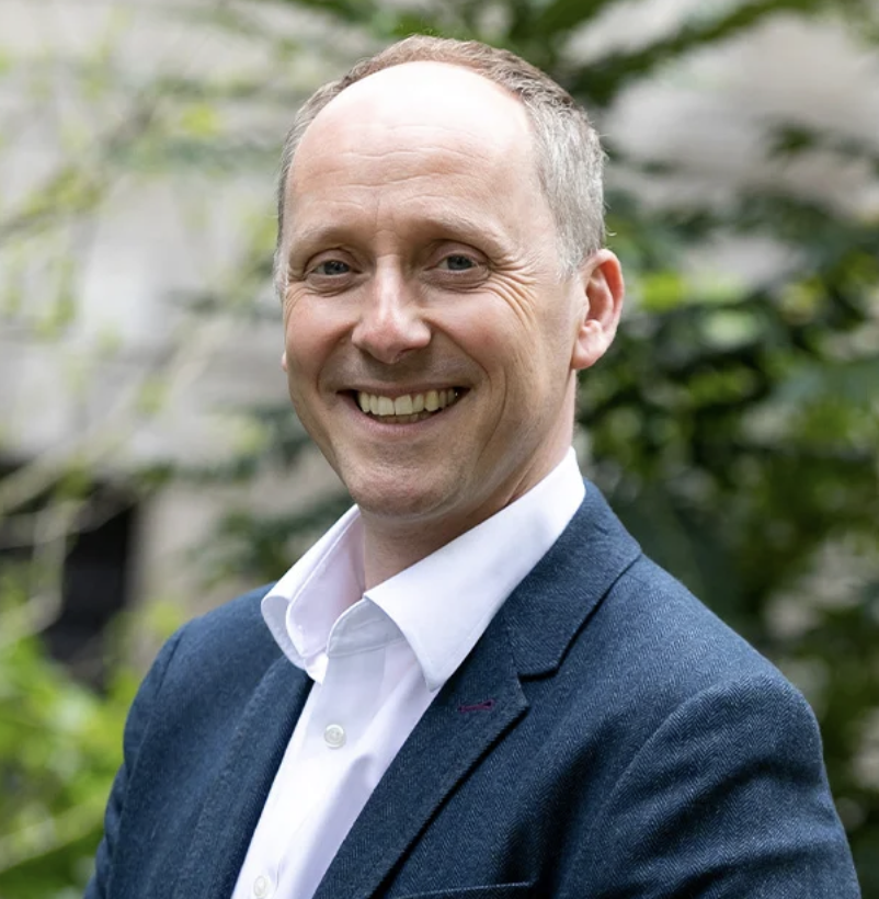 Portrait of a smiling man in a white shirt and navy blazer standing outdoors with greenery in the background.