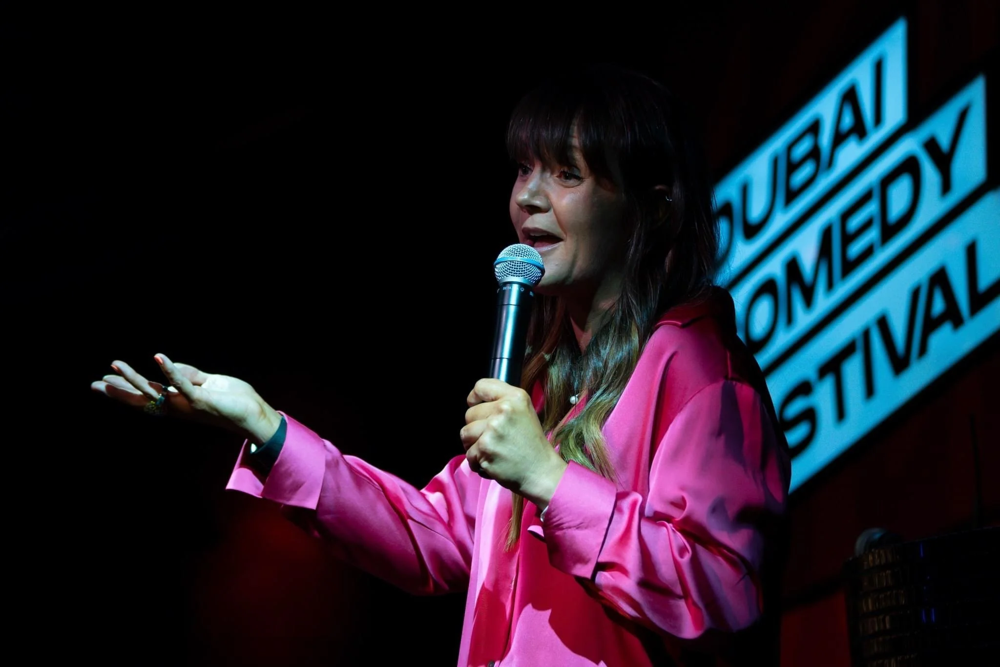 A woman in a pink blouse holds a microphone, performing at the Dubai Comedy Festival.