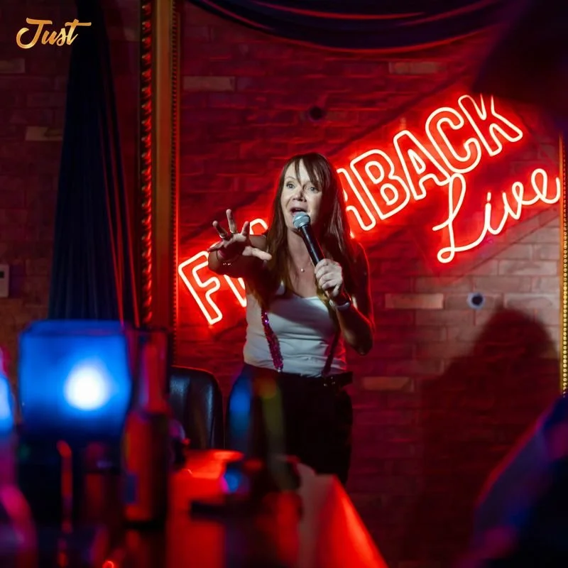 A woman performing comedy on stage with a microphone, in front of a neon sign reading 'Flashback Live'. The setting is a dimly-lit club or bar with a brick wall backdrop and various bottles on a table in the foreground.