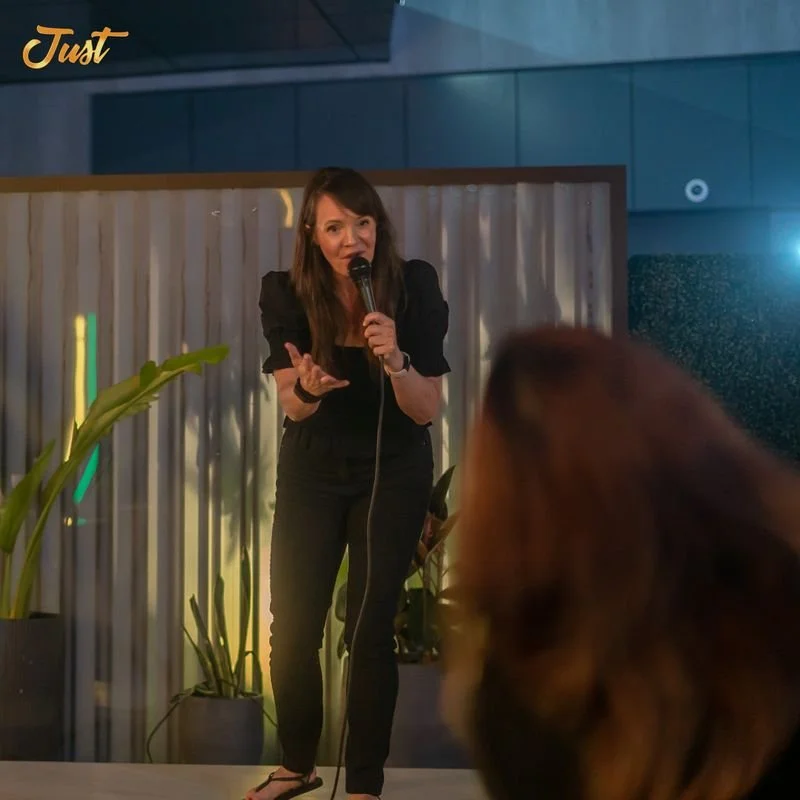Woman performing stand-up comedy on stage, holding a microphone, with audience in the foreground and plants in the background.