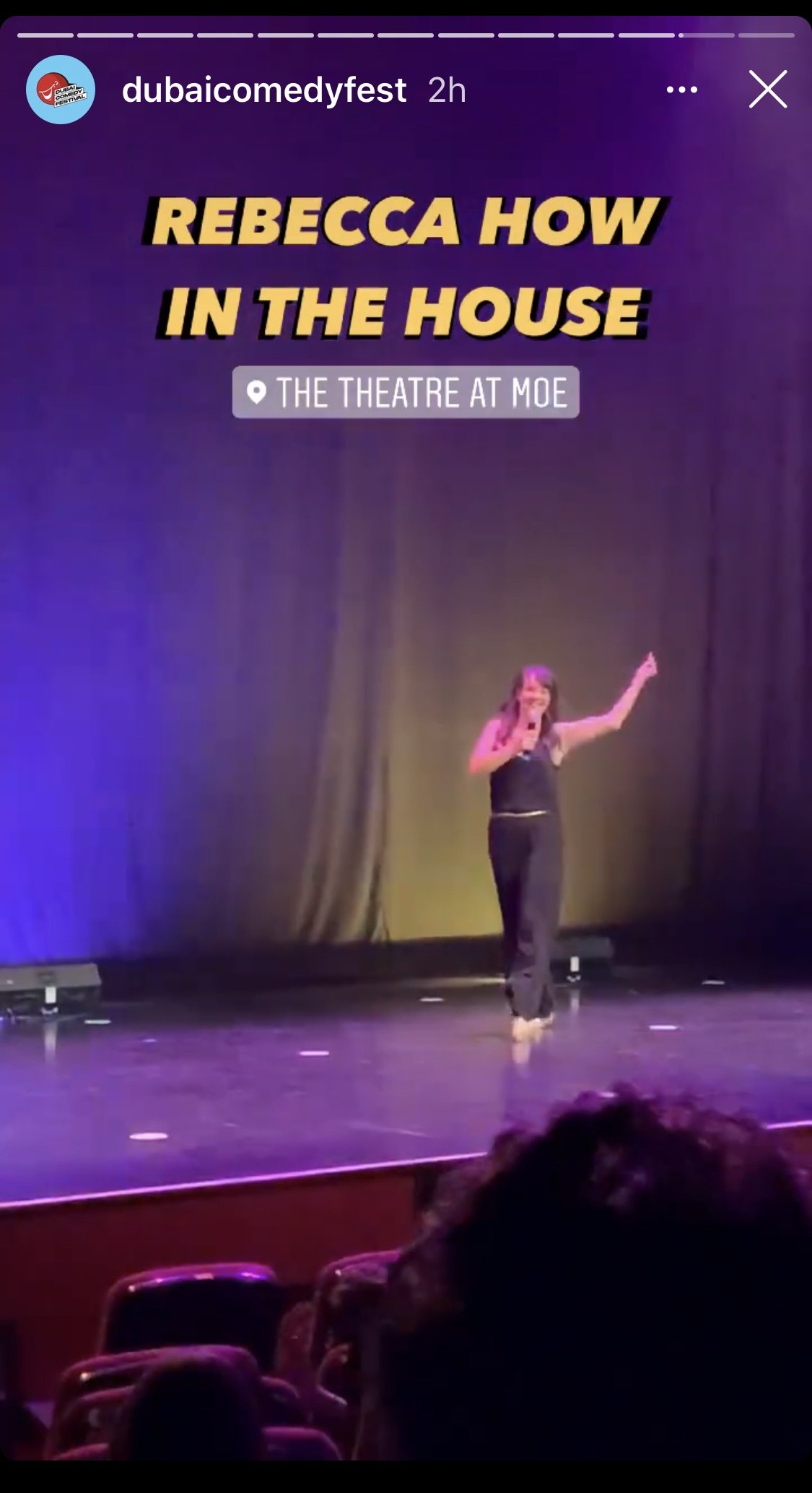 Comedian on stage at Dubai Comedy Festival in theater, wearing black outfit, holding a microphone, with audience seated in foreground; text overlay 'Rebecca How in the House' and 'The Theatre at MOE.'