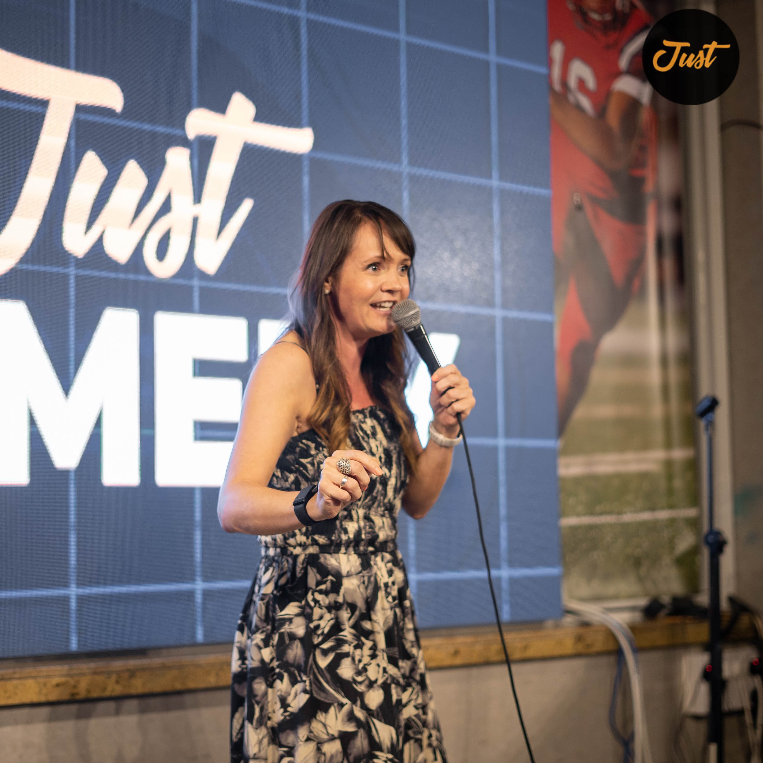 Woman performing stand-up comedy on stage with a microphone, in front of a backdrop displaying the words 'Just Comedy'.
