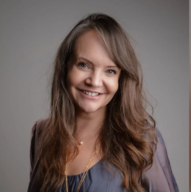 A woman with long wavy brown hair smiling at the camera, wearing a sheer purple blouse and a gold necklace with a pearl pendant.