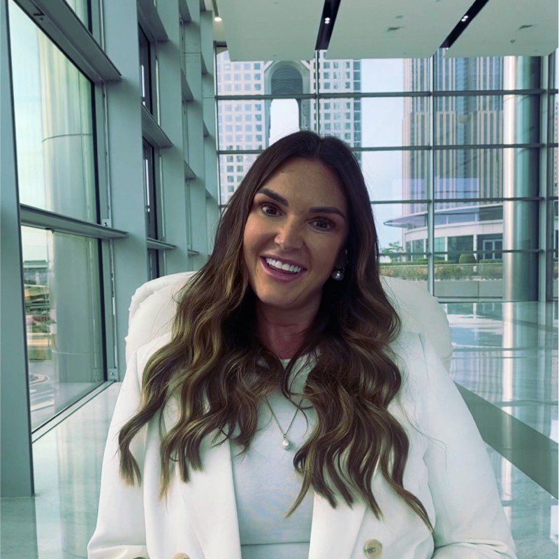 A smiling woman with long wavy hair, wearing a white blazer and pearl earrings, sitting in a modern glass building with city skyscrapers in the background.