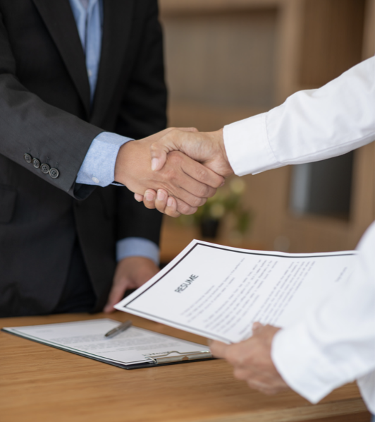 Two people shaking hands over a desk with a contract and pen, in a professional office setting.