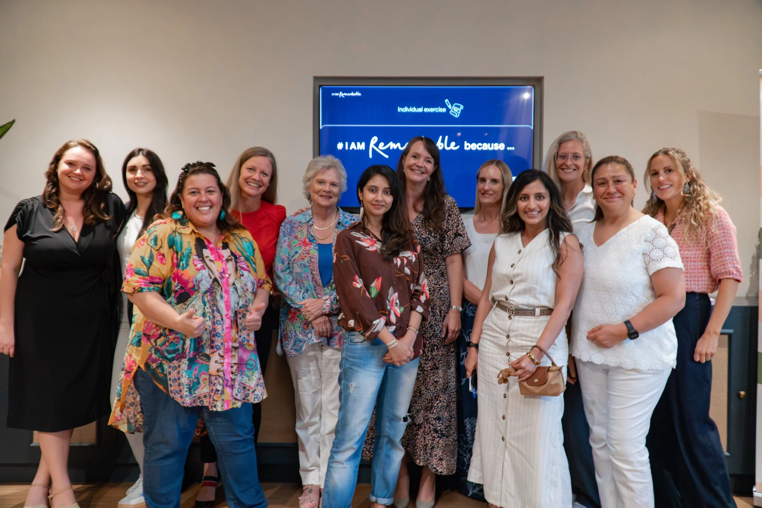 Group of diverse women standing together indoors, smiling at the camera, with a blue screen behind them that says "Individual exercise" and "I am Remarkable because...".