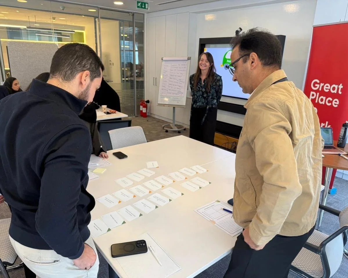 Two men stand at a conference table looking at arranged cards, with a woman smiling in the background near a whiteboard and a large screen in a modern office setting.