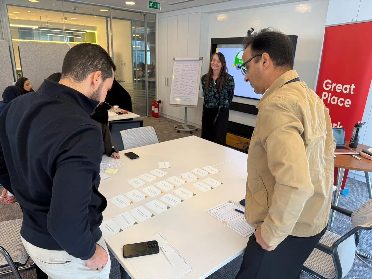 Two men standing at a table in a meeting room, looking at arranged cards on the table, with a woman smiling in the background near a whiteboard and screen, in a modern office space.