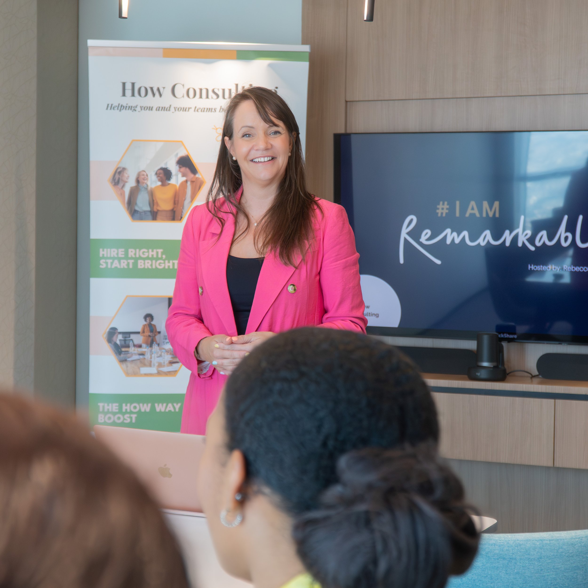 A woman in a pink blazer giving a presentation in a conference room, with a large screen behind her displaying a hashtag and the word "Remarkable."