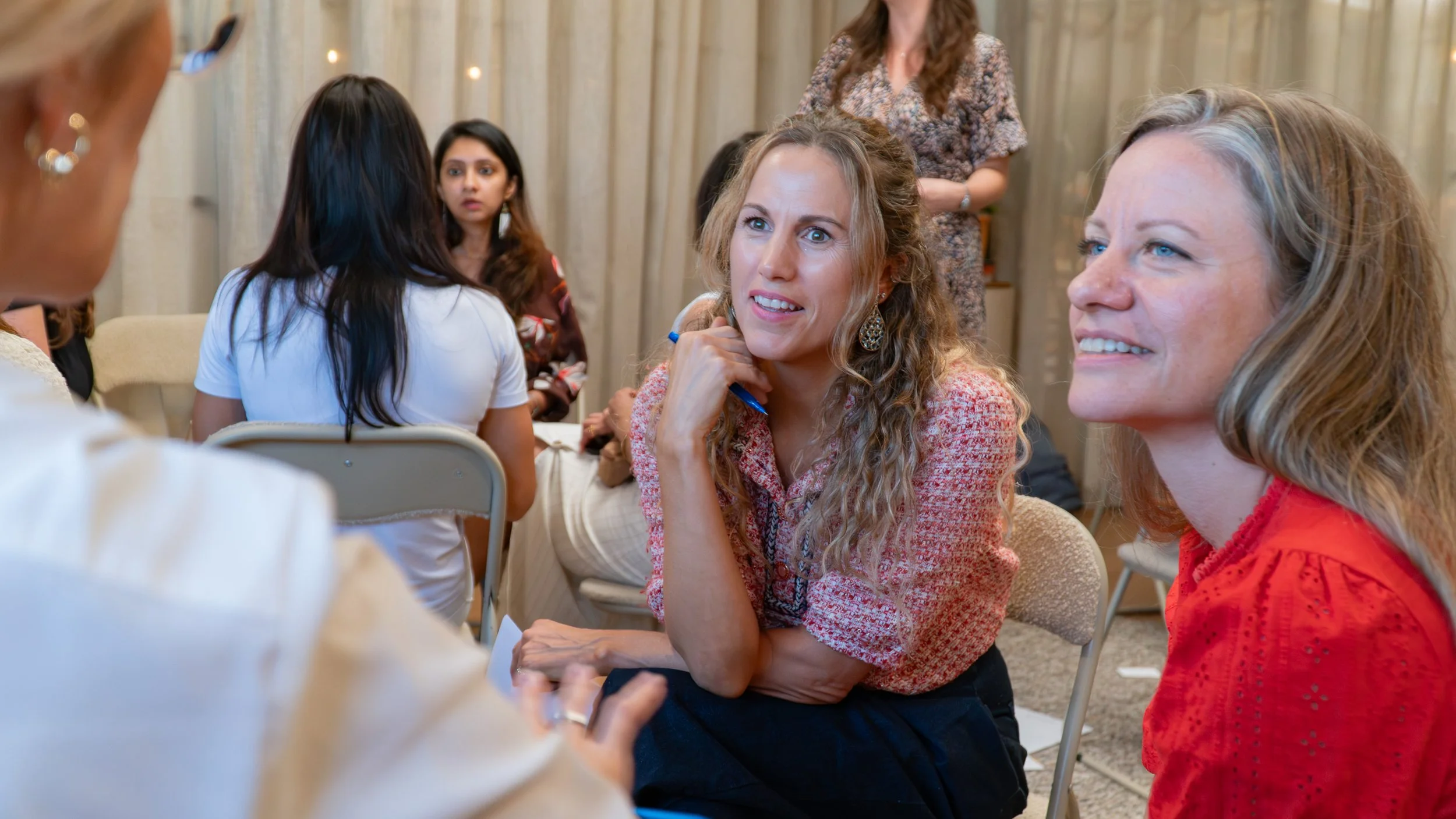 Group of women engaged in conversation at a social gathering, seated in a circle, with one woman smiling and another listening attentively.