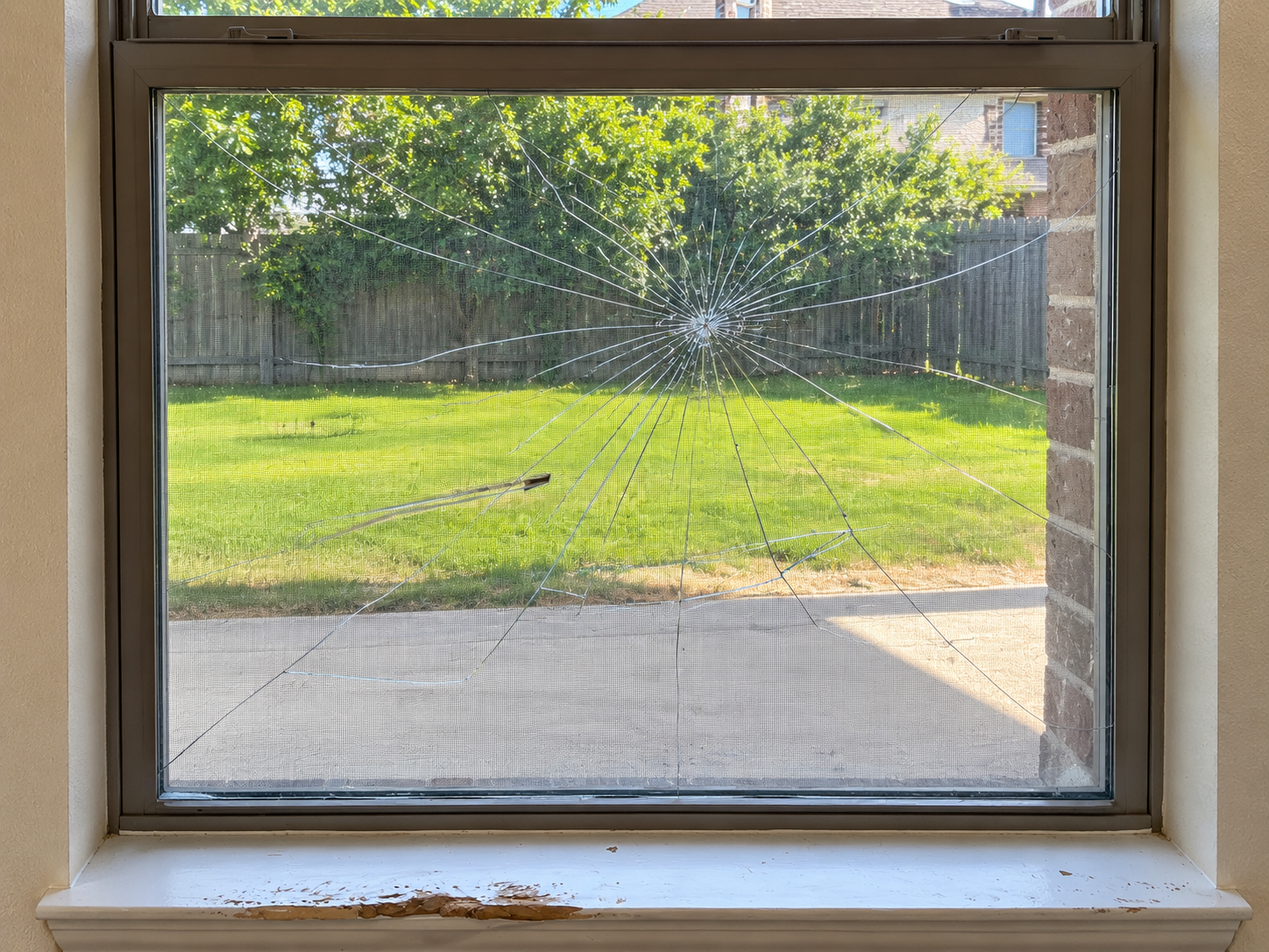 Broken window with a large spiderweb crack, view of a backyard with green grass, a wooden fence, and trees in the background.