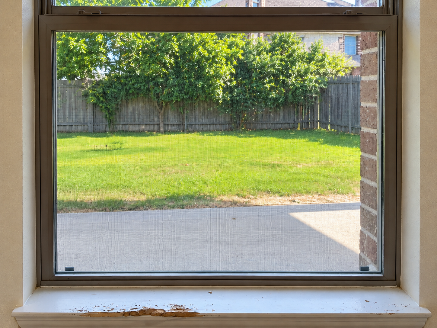 View of a backyard with green grass, trees, and a wooden fence, seen through a window with a brick wall on the right side.