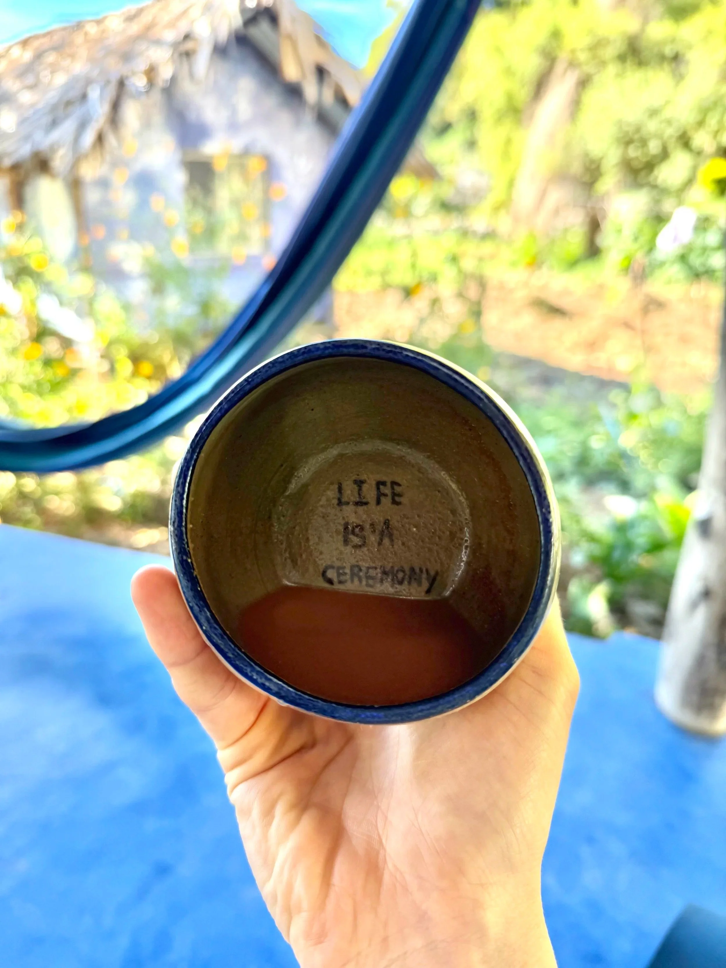 Hand holding a ceramic cup with the words 'LIFE IS A CEREMONY' written inside, against a background of a garden and a wooden structure.