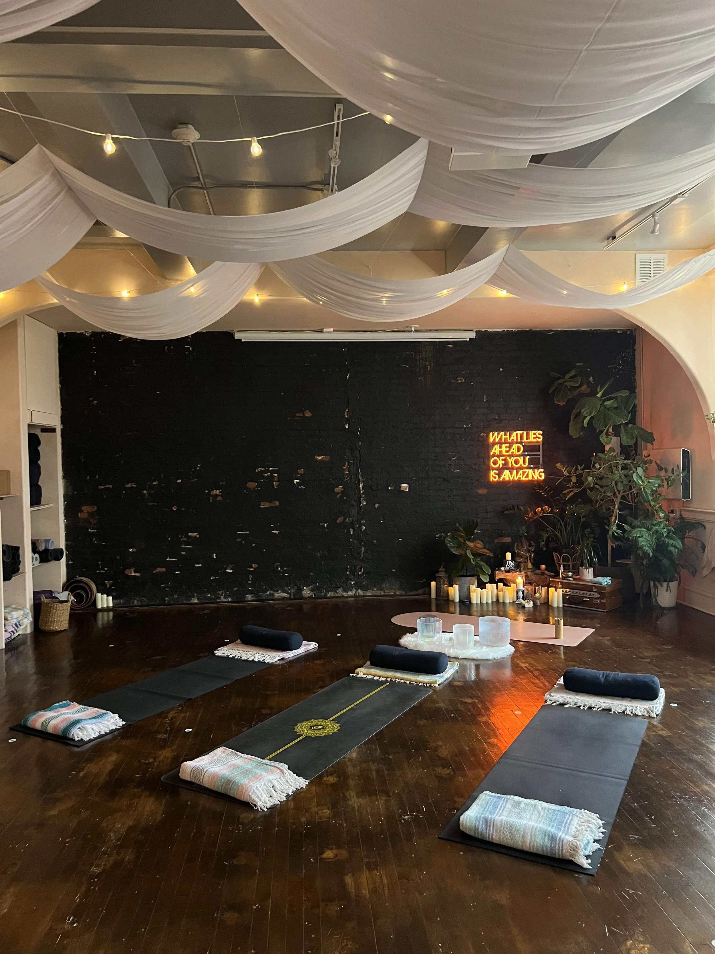A yoga studio with three black mats, each with a rolled towel and long bolster cushion, on a wooden floor. The ceiling is decorated with white fabric drapes and string lights. The back wall has a black brick area with a neon sign that reads, 'What lies ahead of you is amazing.' Candles and plants are arranged along the wall near the sign.