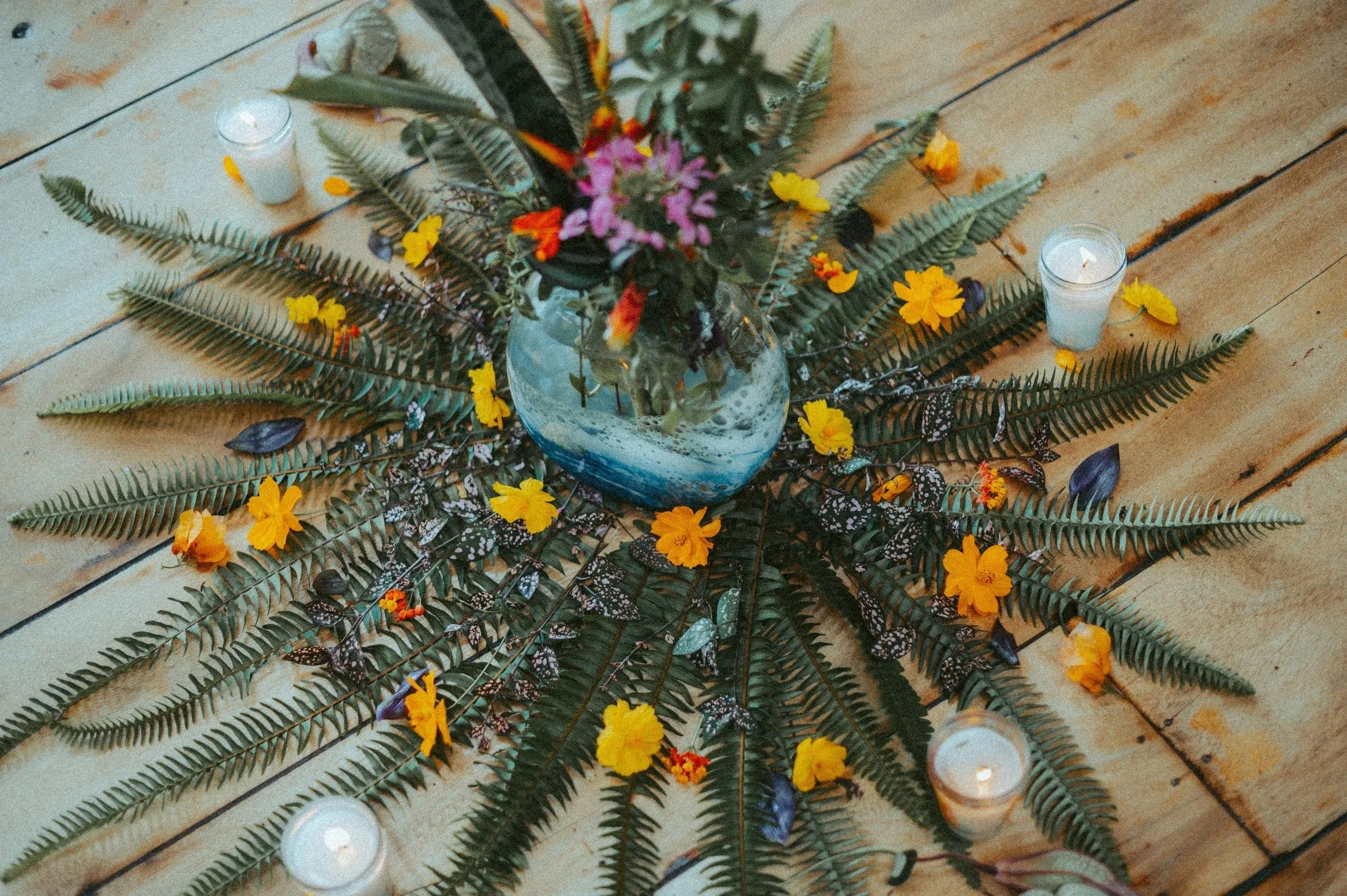 A floral arrangement on a wooden floor with a blue vase containing flowers, surrounded by large green fern leaves, yellow and orange flowers, small butterflies, and four lit candles in glass holders.
