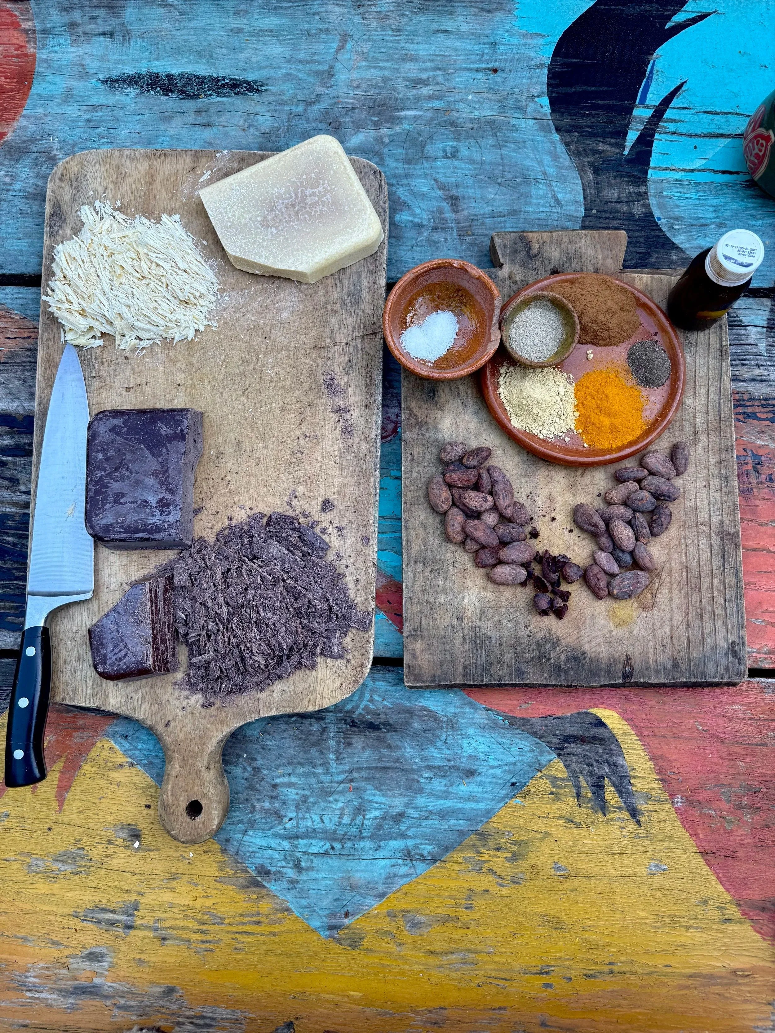 Cutting board with cheese, shredded meat, and blocks of meat, alongside a knife, on a colorful, weathered wooden table. On a separate smaller wooden board, there are various spices, nuts, and a small bottle.