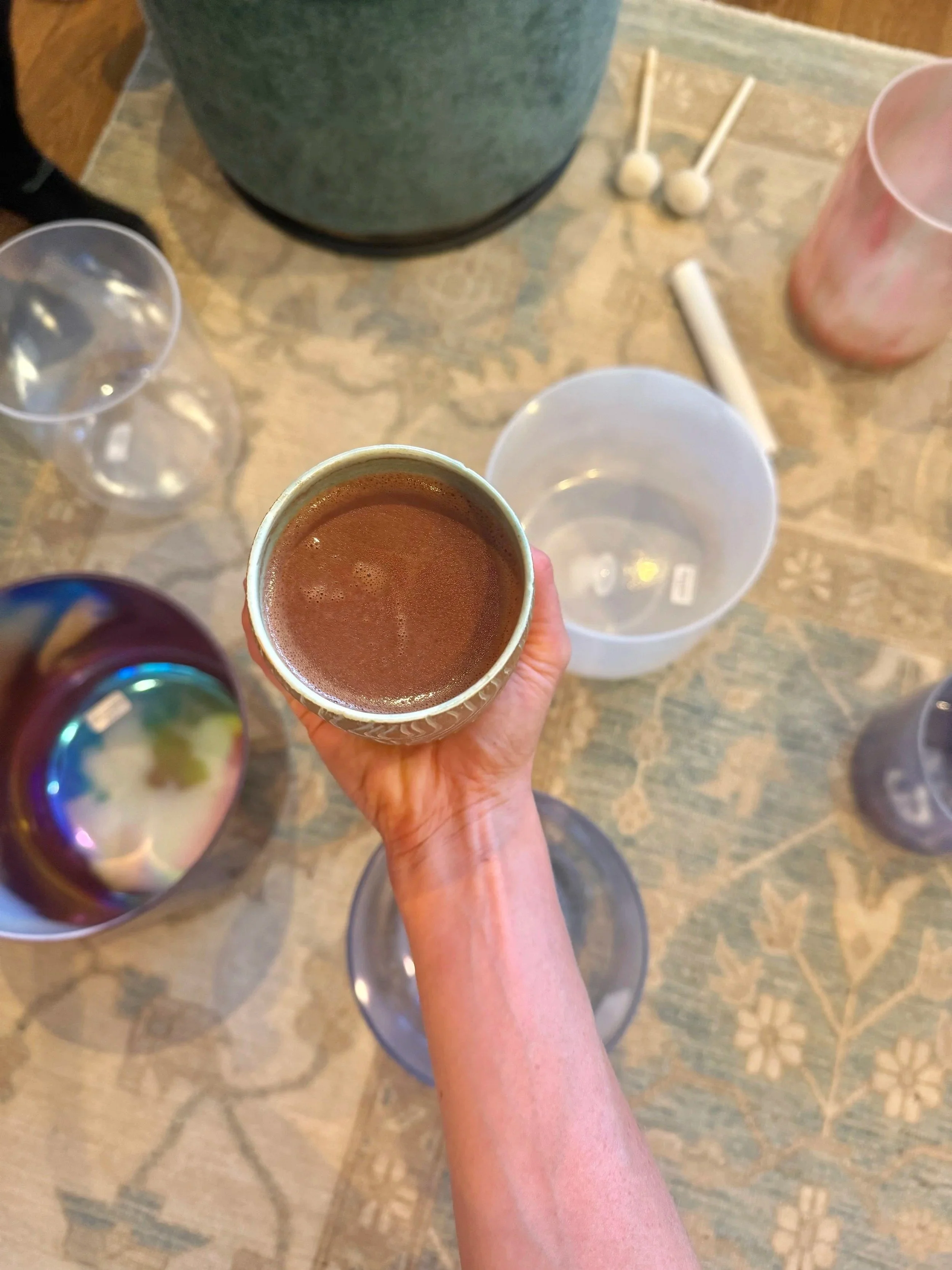 A person holding a cup of hot chocolate over a table with empty glasses, a pink cup, a white bowl, and various candles or decorative items.