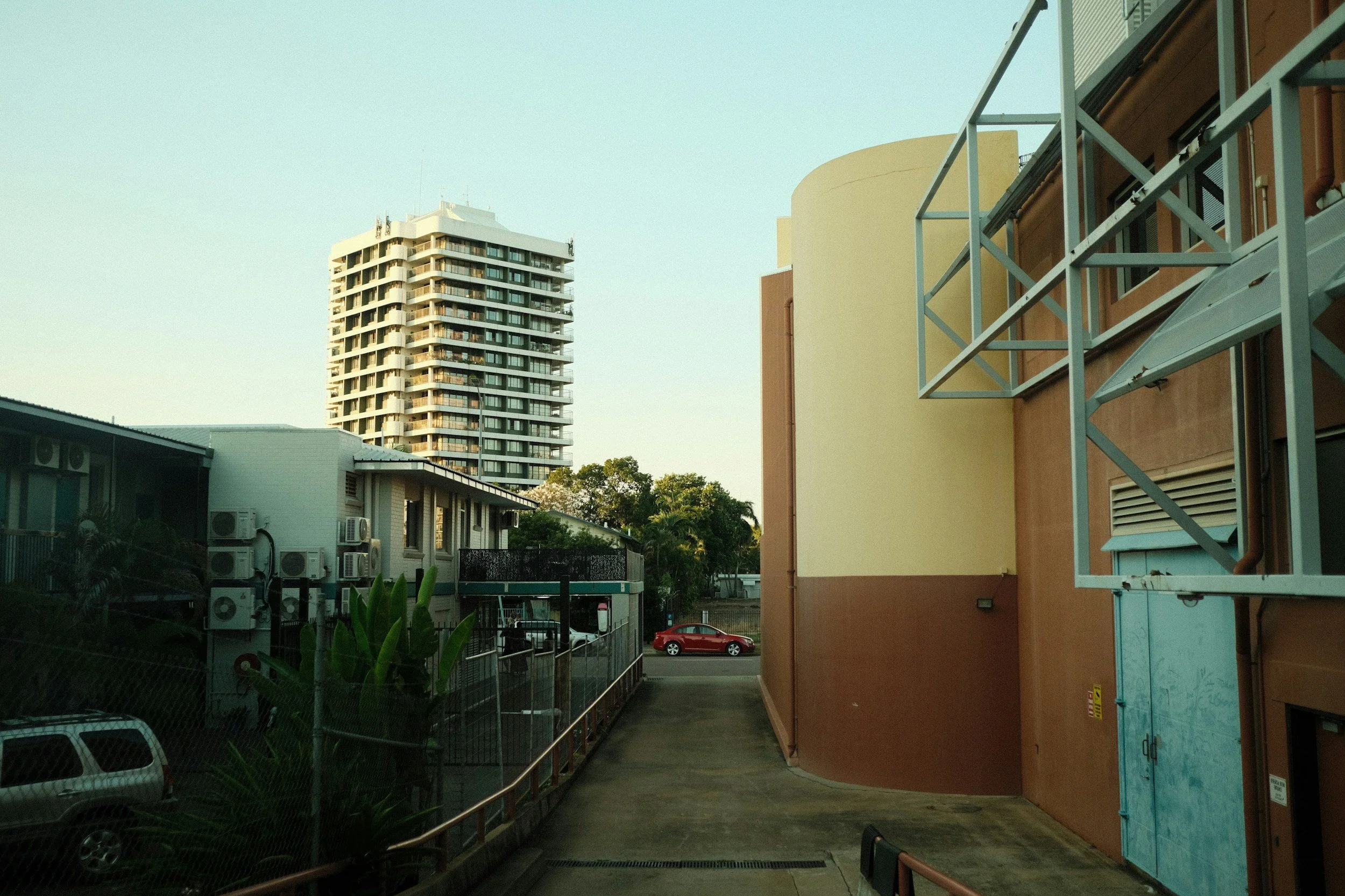 View of a cityscape with a tall residential building in the background and a brown apartment building on the right, with air conditioning units on the side. A parking lot with cars and some greenery are also visible.