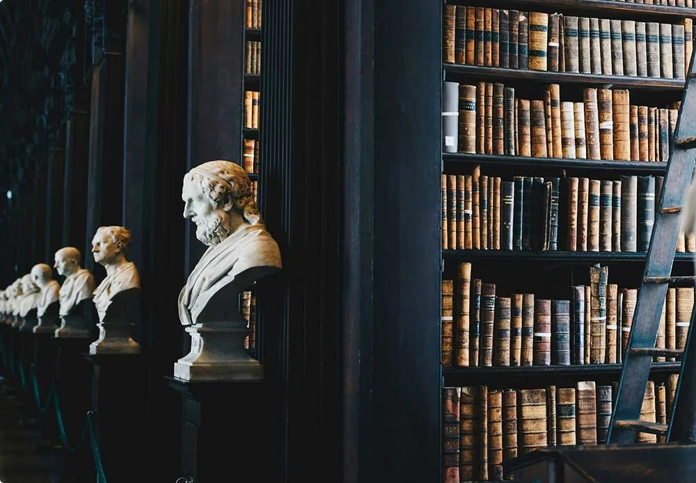 A row of white marble bust sculptures of famous figures in front of large wooden bookshelves filled with old books in a historical library.