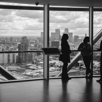 Two people standing inside a high-rise building looking out at a cityscape through large floor-to-ceiling windows, with a display stand in the foreground.