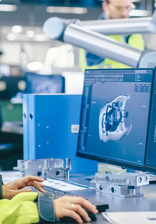 A person working on a computer designing a 3D model of a mechanical part in an engineering or manufacturing facility.