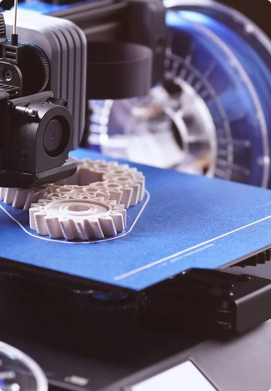 A 3D printer creating white gears on a blue printing bed, with the printer's nozzle and part of the machinery visible.