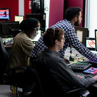 Three men working at computer stations in a tech office, with pink walls and multiple monitors.