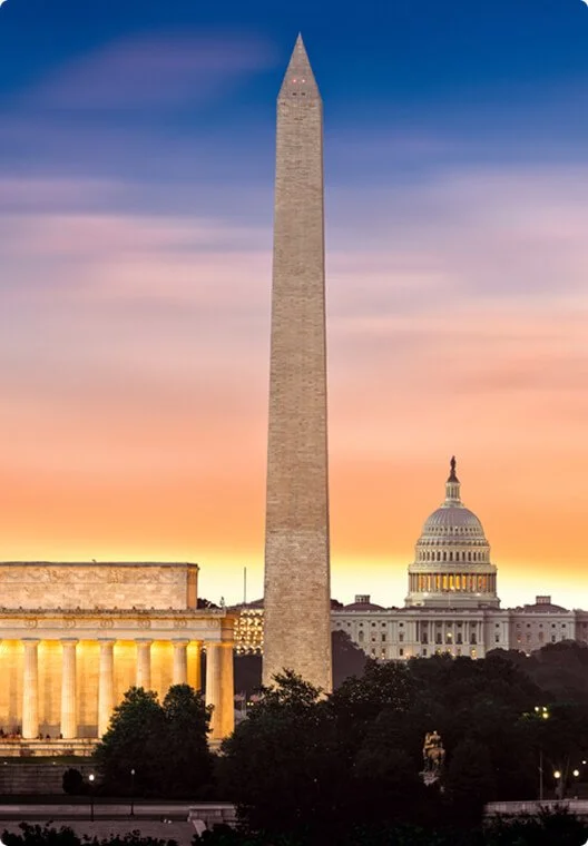 A large obelisk monument with the U.S. Capitol building in the background during sunset, with a colorful sky.