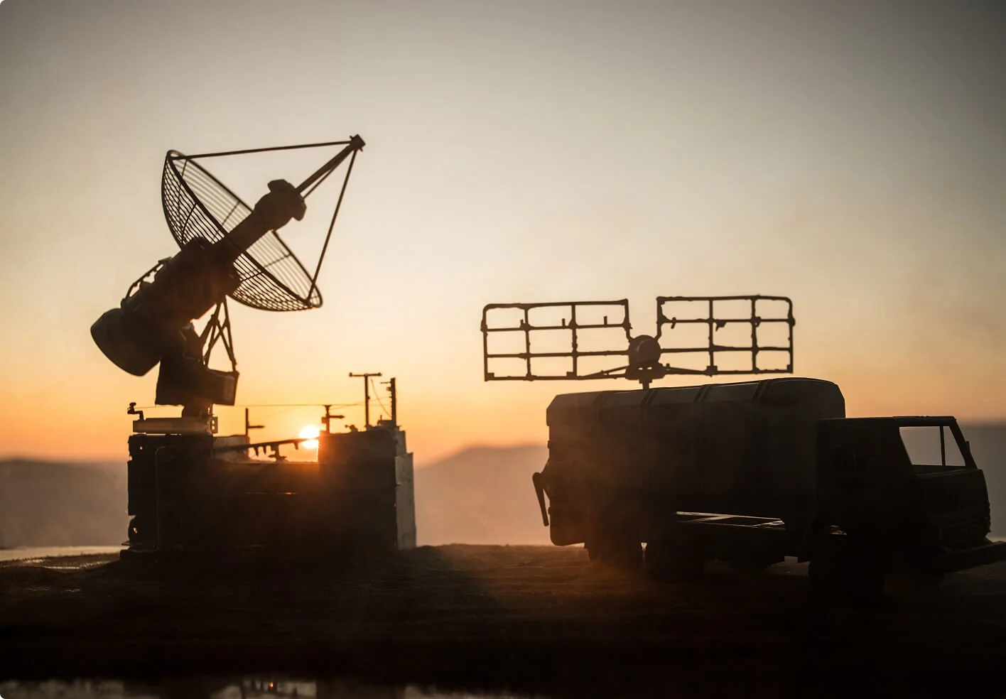 Silhouettes of satellite dishes and communication equipment on trucks against a sunset sky.