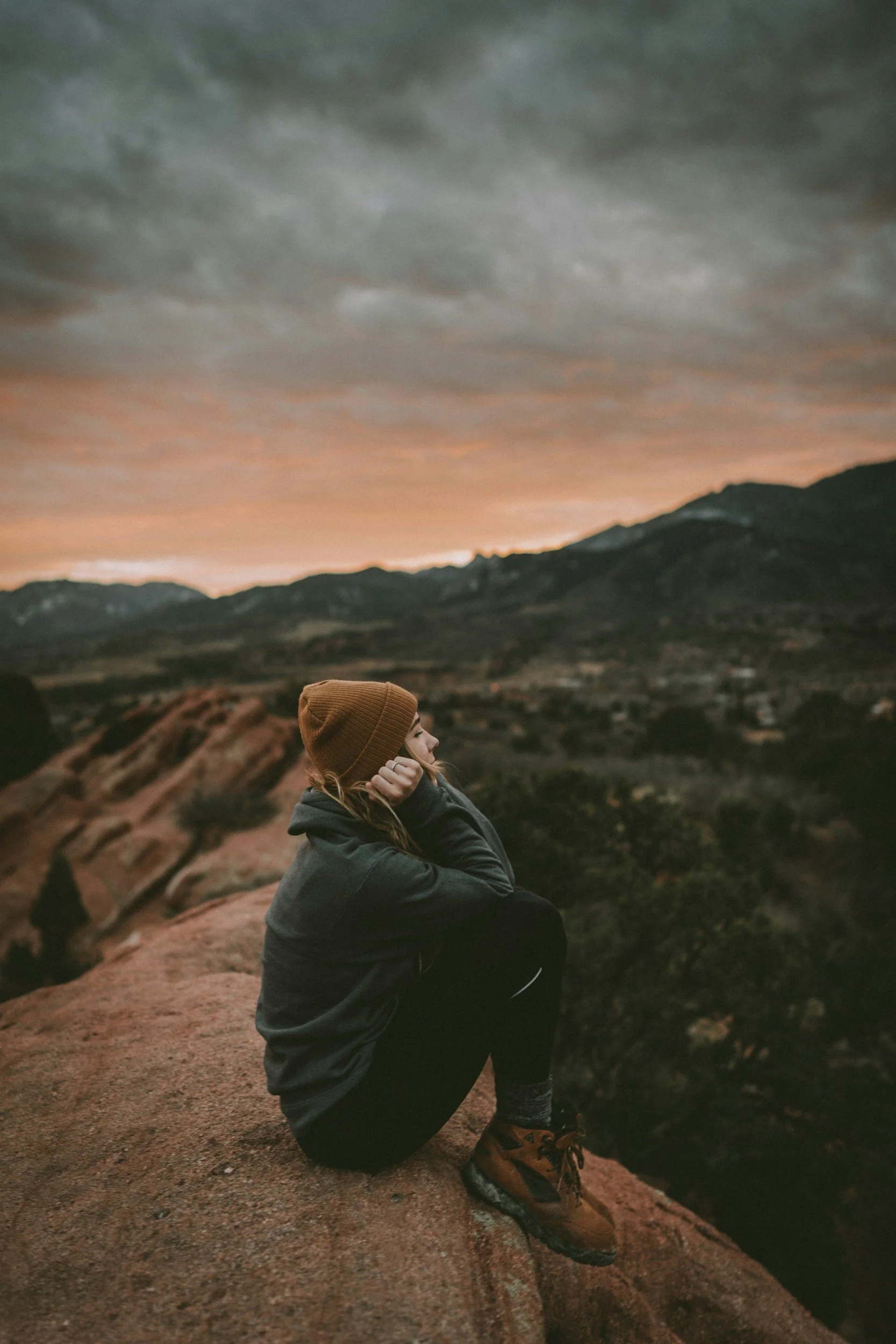 A person sitting on a rocky ledge, looking thoughtfully at a mountain landscape with a dramatic sky during sunset.