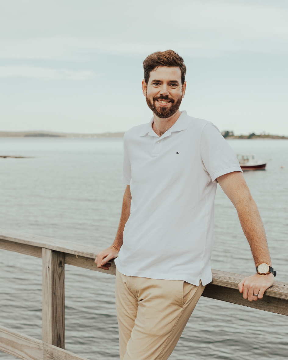 Man with brown hair and beard in white polo shirt and beige pants leaning on a wooden dock railing by a body of water with a boat in the background.