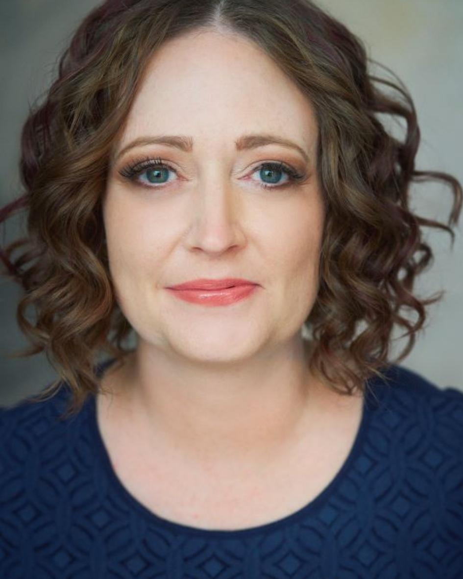 Close-up of a woman with light skin, curly brown hair, blue eyes, light makeup, and a gentle smile, wearing a navy blue textured top.
