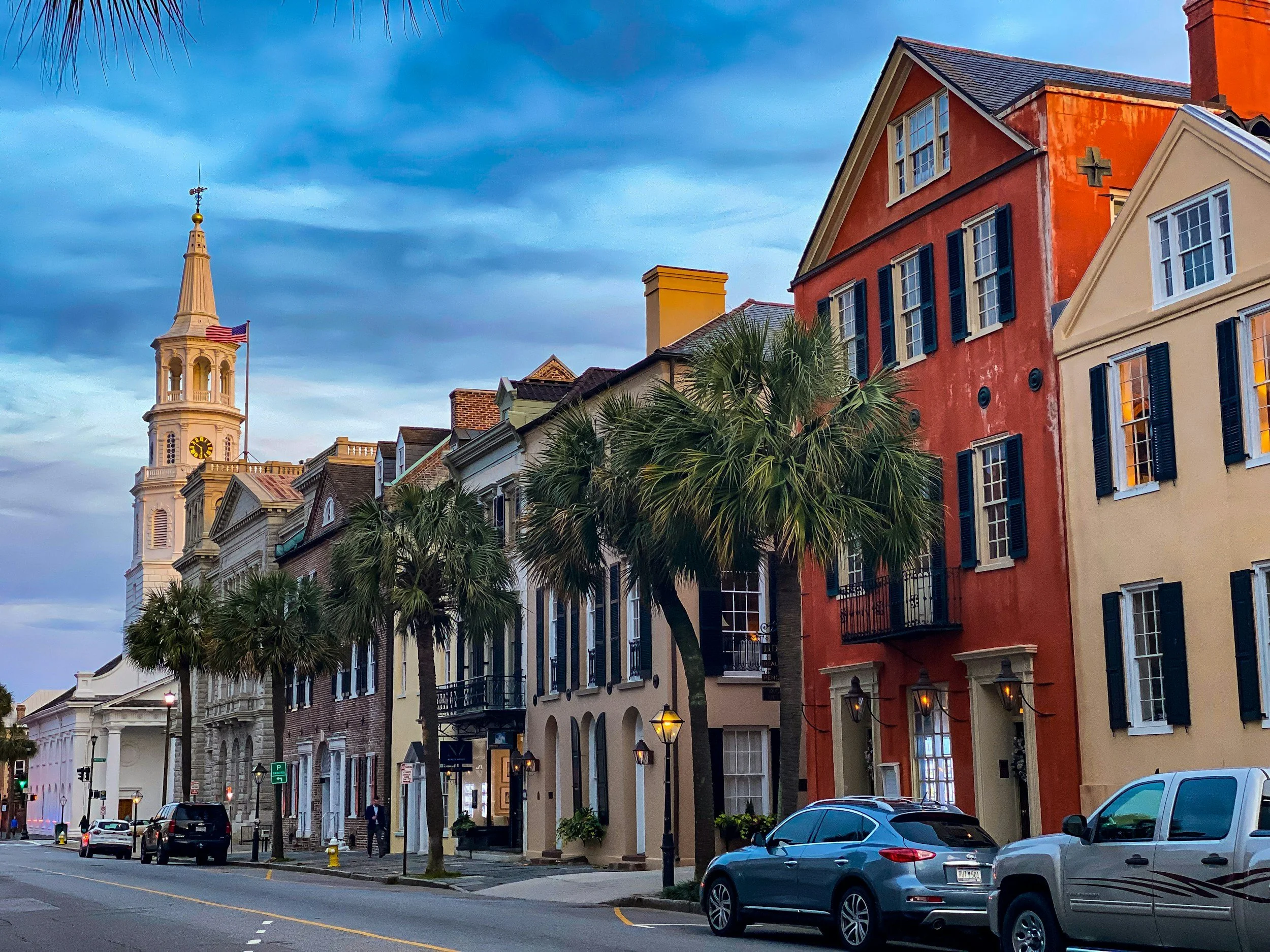 Colorful buildings on a city street with palm trees, parked cars, and a clock tower in the background under a cloudy sky.