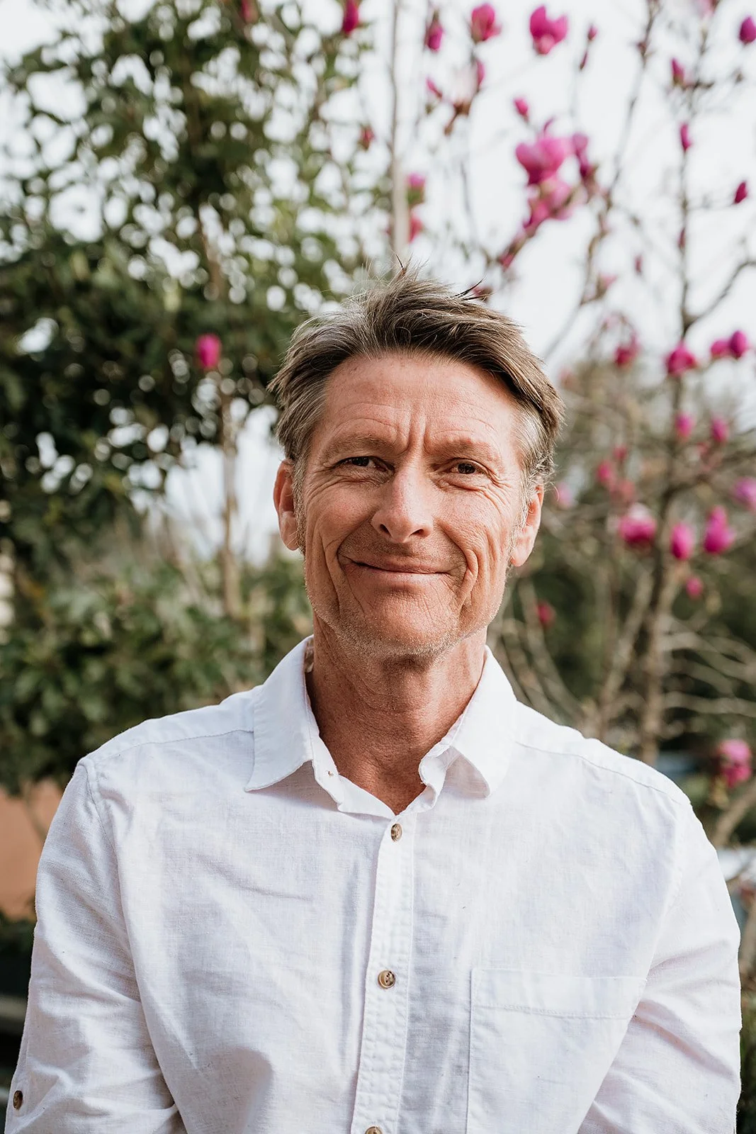 A middle-aged man with gray hair and a slight smile, wearing a white button-up shirt, standing outdoors with pink flowering trees in the background.