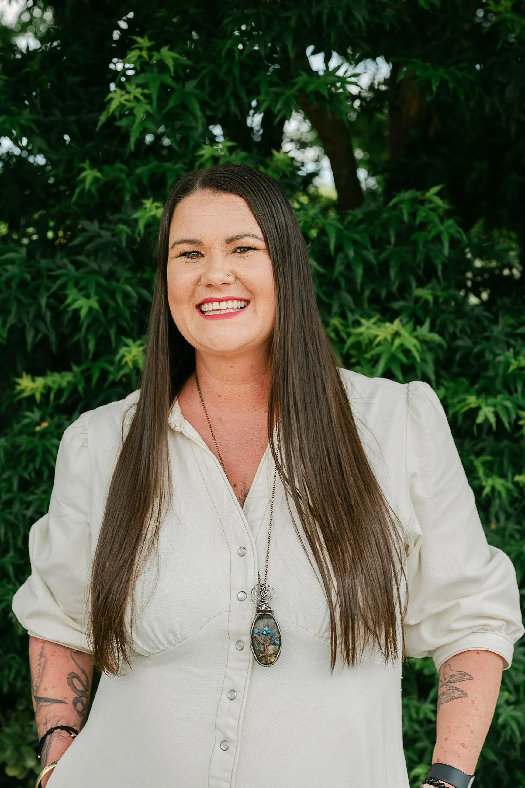 A smiling woman with long brown hair, wearing a light-colored blouse and a large statement necklace, standing outdoors in front of green foliage.